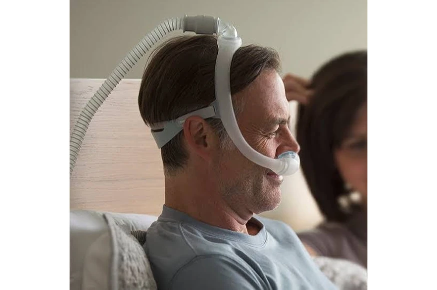 Man wearing Philips DreamWear Gel Pillow nasal mask, sitting up in bed with partner asleep beside him