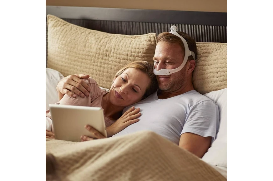Couple in bed, man wearing Philips DreamWear nasal mask, woman using a tablet beside him