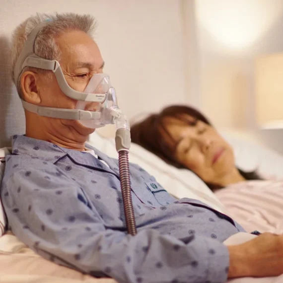 Man wearing ResMed AirFit F20 mask sitting up in bed with partner asleep beside him