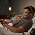 Man wearing ResMed AirFit F20 mask using a tablet in bed with partner asleep beside him